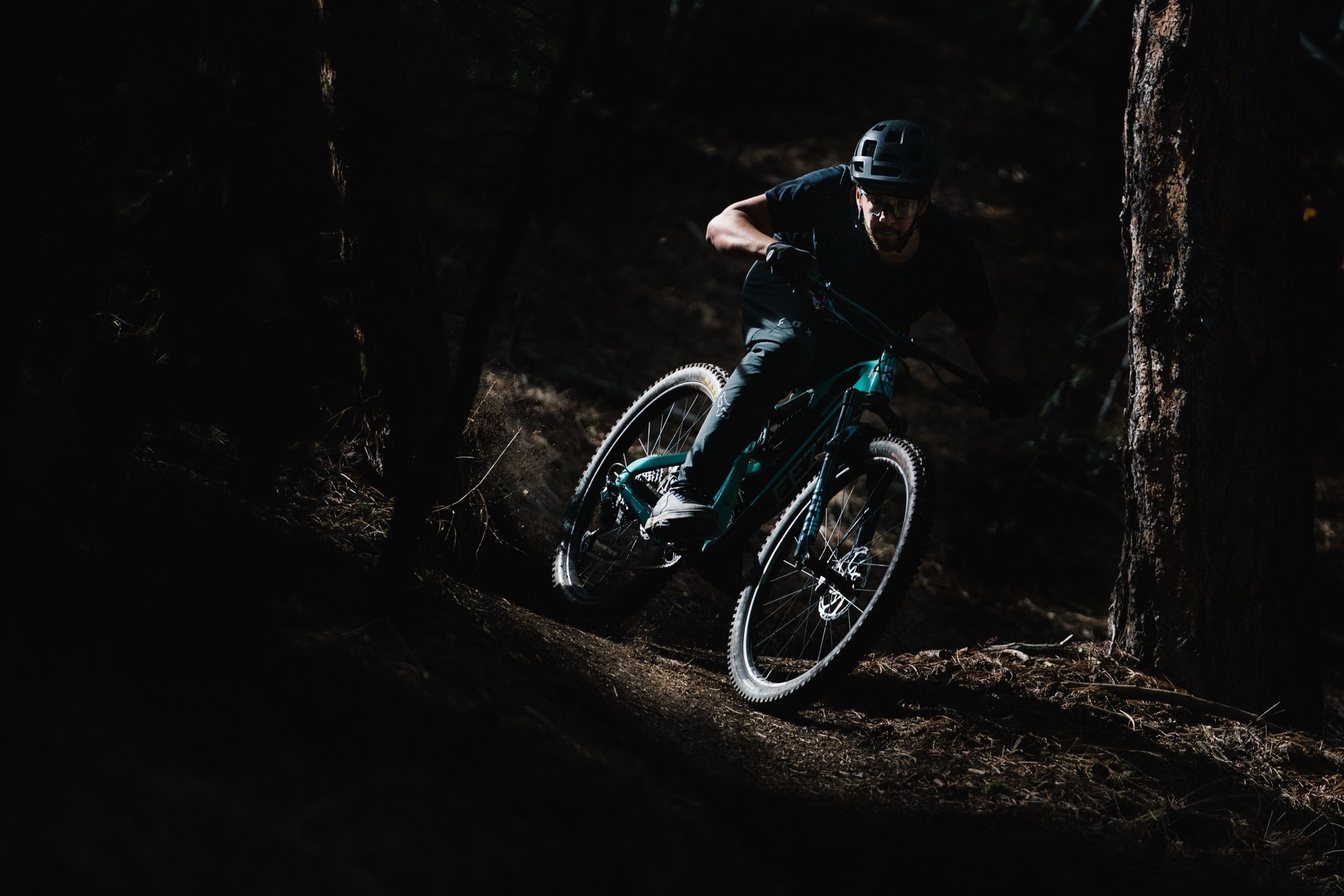 A mountain biker cornering hard on a dusty forest trail, lit by a shaft of sunlight